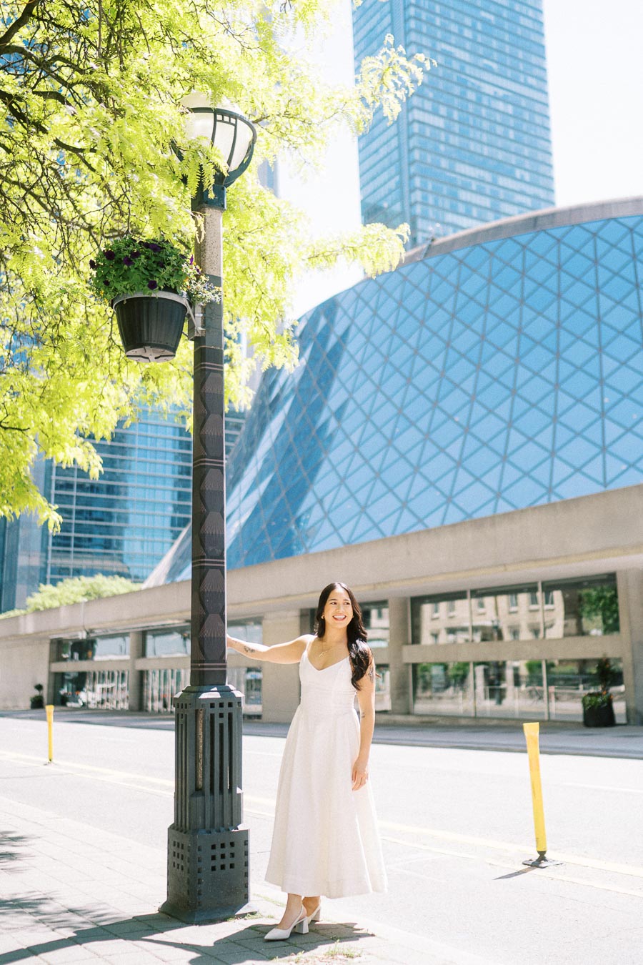 A woman in a white dress stands beside a lamppost on a city sidewalk in downtown Toronto, with glass buildings and greenery visible.