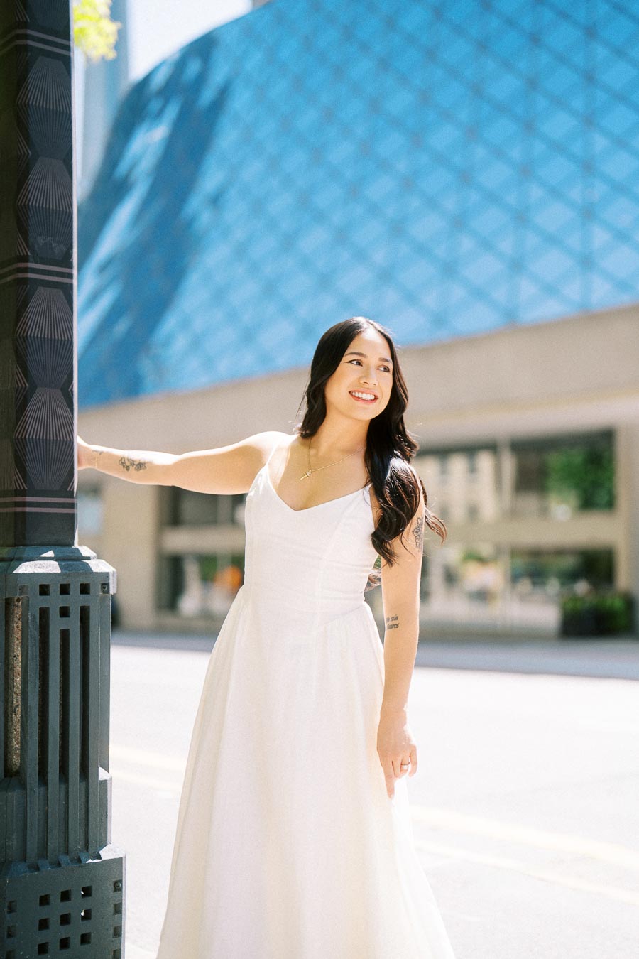 A woman in a white dress smiles while leaning on a lamppost in downtown Toronto with a modern glass building in the background.