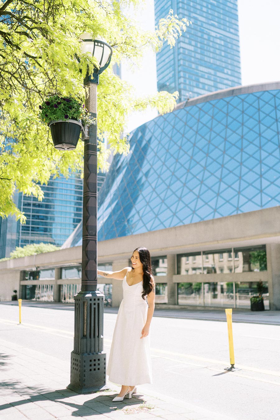 A woman in a white dress stands beside a lamp post on a city sidewalk with a modern glass building behind her in downtown Toronto.