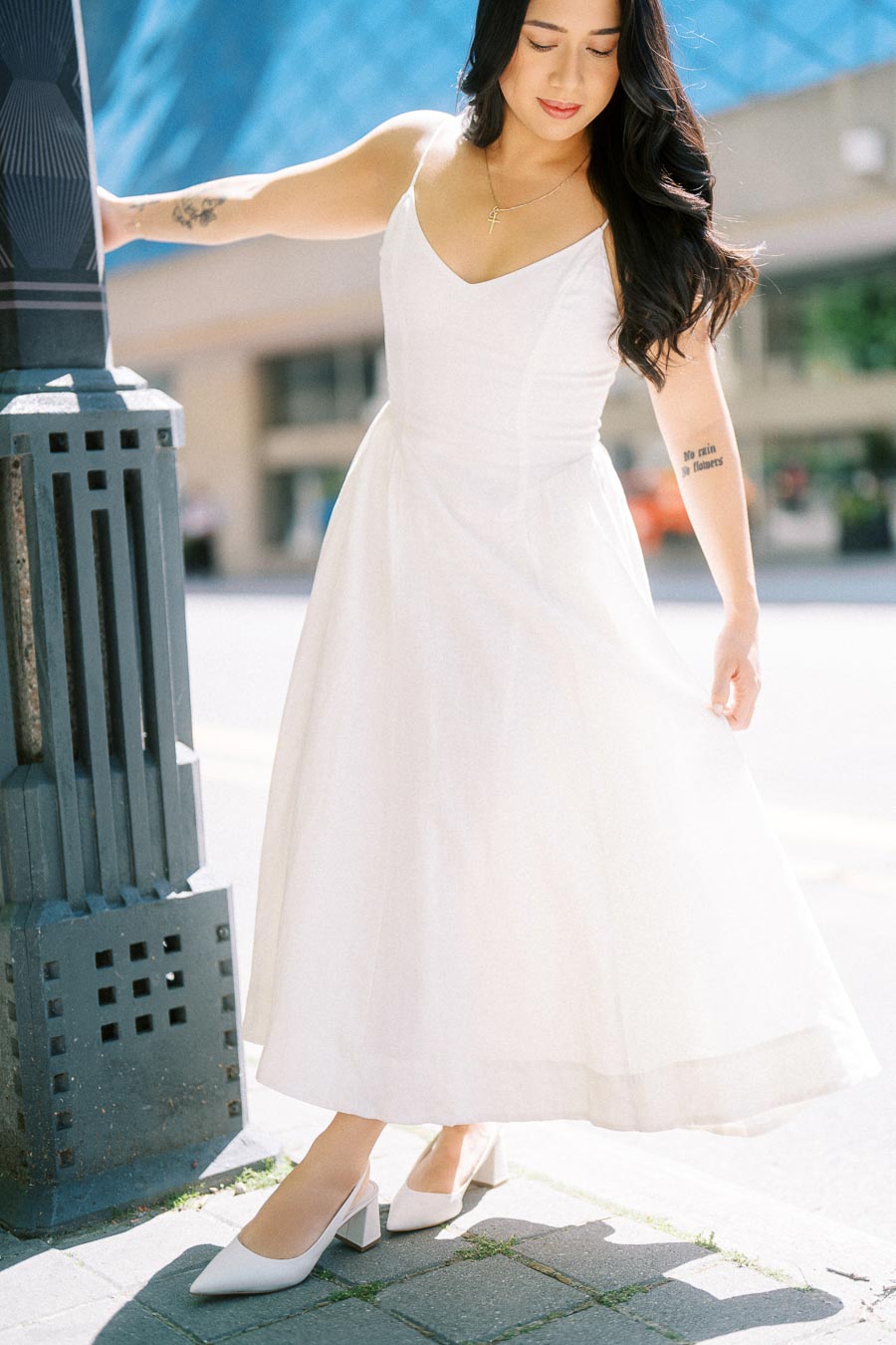 A woman in a white dress stands beside a lamp post on a city sidewalk in downtown Toronto, holding her skirt and looking down.