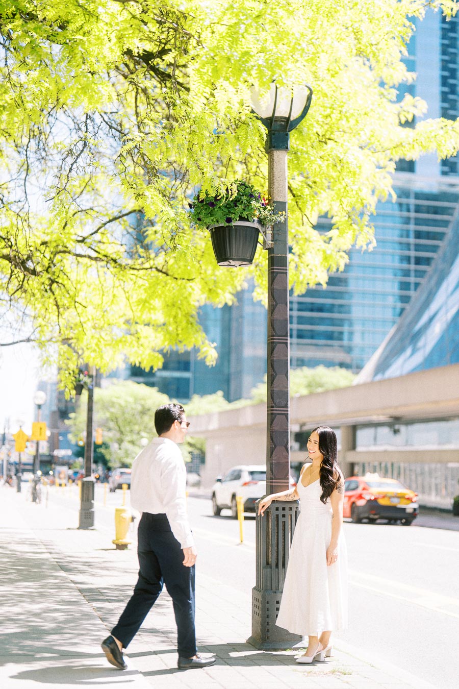 Cassie and Gabriel stand and smile at each other on a sunlit downtown Toronto sidewalk, with green trees and city buildings in the background.