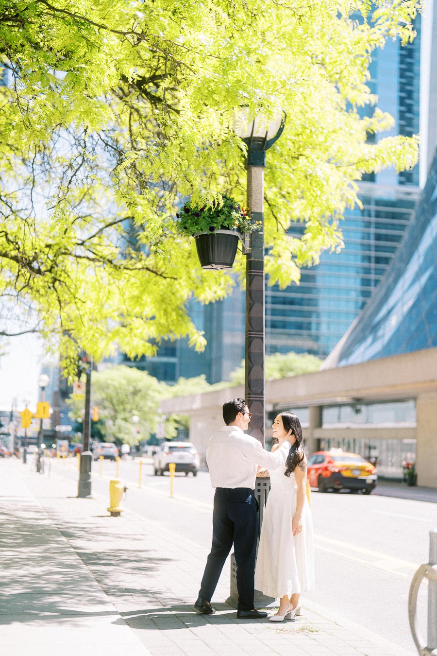 Cassie and Gabriel stand together on a sunlit downtown Toronto street, smiling at each other beneath vibrant green trees and a city lamppost.