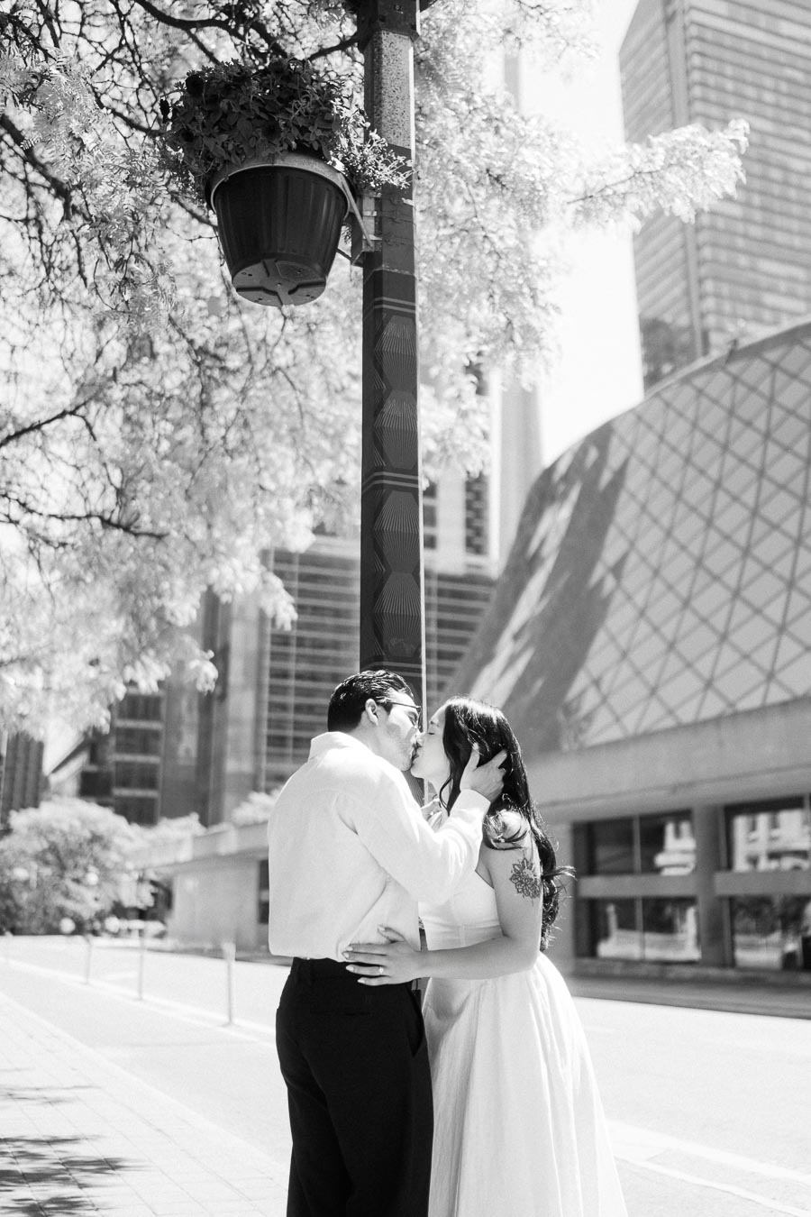 Cassie and Gabriel kissing on a downtown Toronto sidewalk with modern buildings and a tree-lined street in the background.