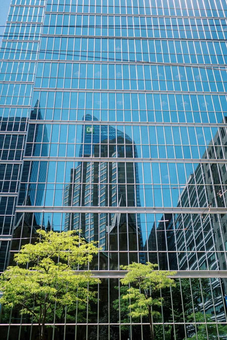 Reflections of Toronto skyscrapers, historic church architecture, and green trees in the glass windows of a downtown office building.