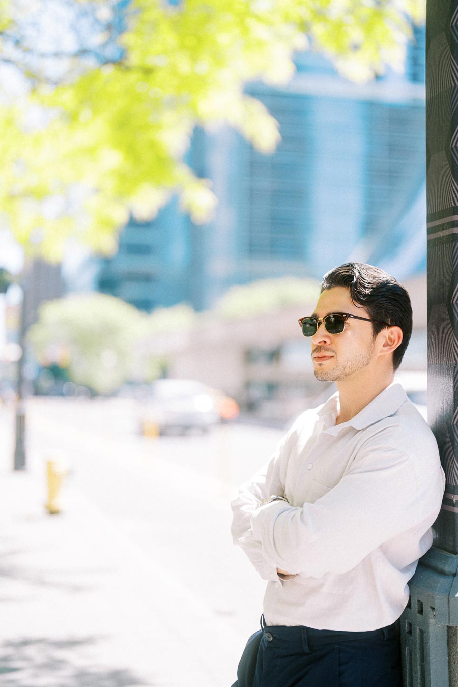 Gabriel wearing sunglasses and a white shirt, leaning against a lamp post on a sunny downtown Toronto street with trees and modern buildings in the background.