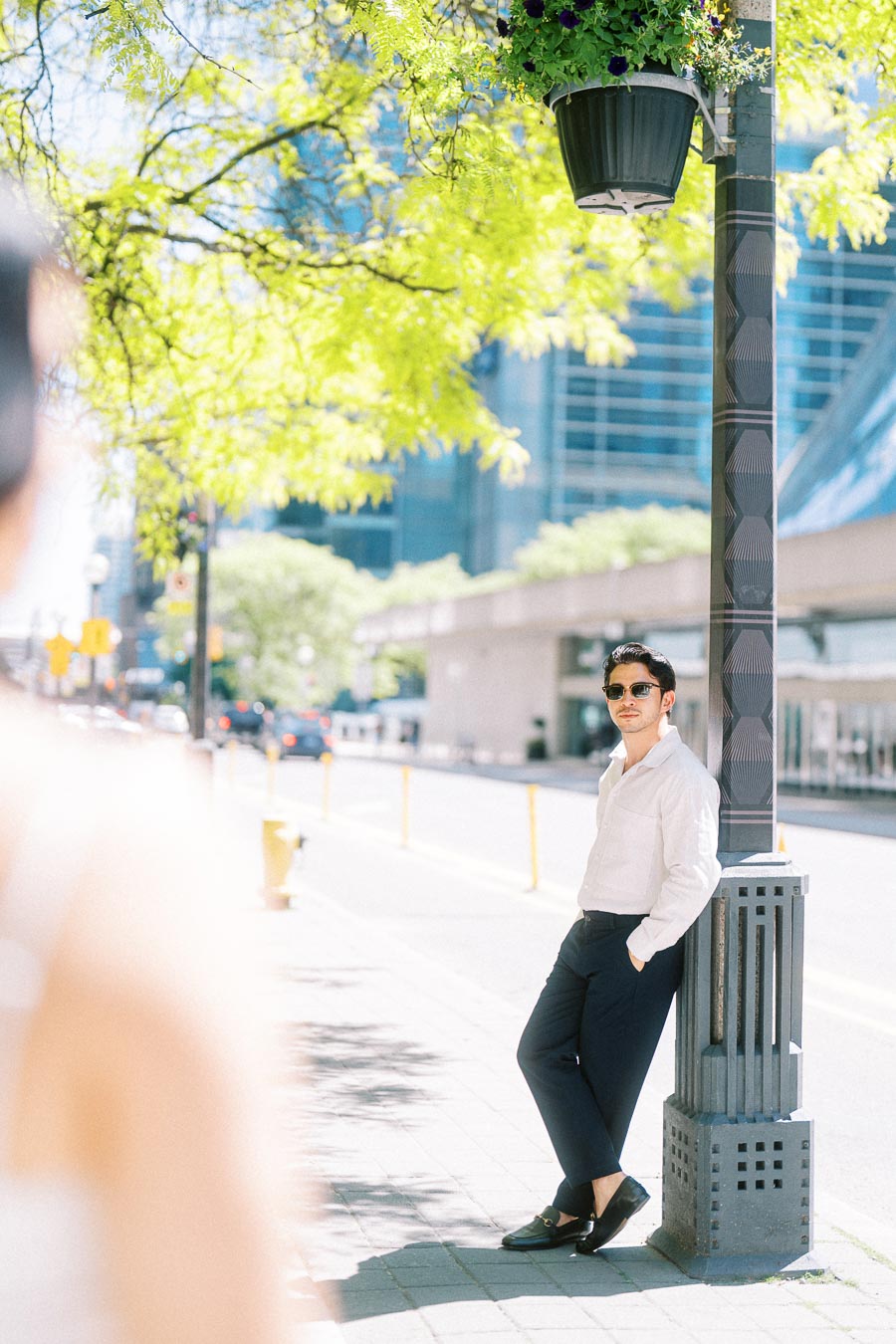 Gabriel leaning against a lamp post on a bright city sidewalk in downtown Toronto, with Cassie slightly blurred in the foreground.