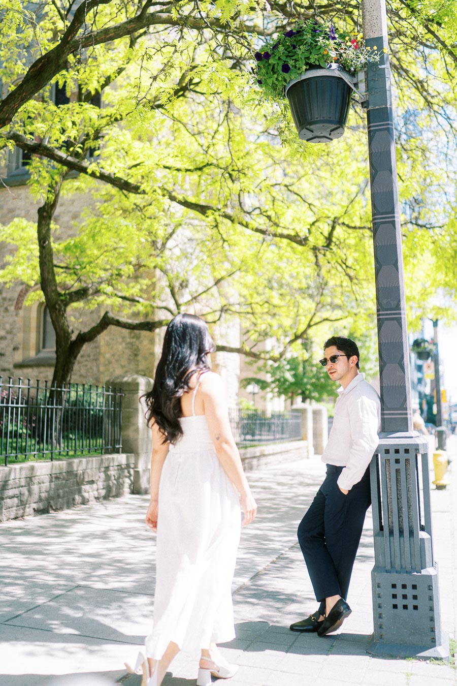 Cassie and Gabriel enjoying a sunny engagement session on a tree-lined sidewalk in downtown Toronto, with historic architecture in the background.