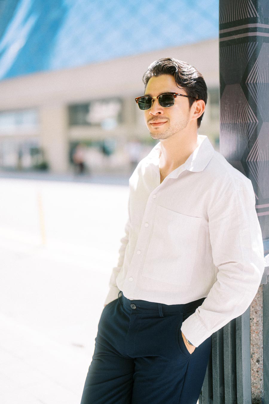 Gabriel stands outdoors in downtown Toronto, leaning against a patterned column and wearing sunglasses and a white shirt.