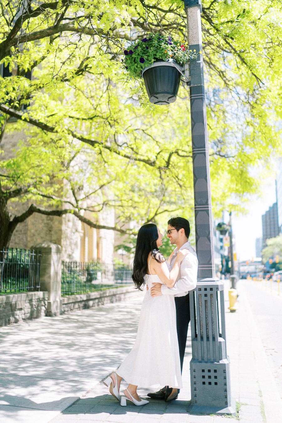 Cassie and Gabriel embrace and smile at each other under a streetlamp with flowers, surrounded by bright green trees in downtown Toronto.