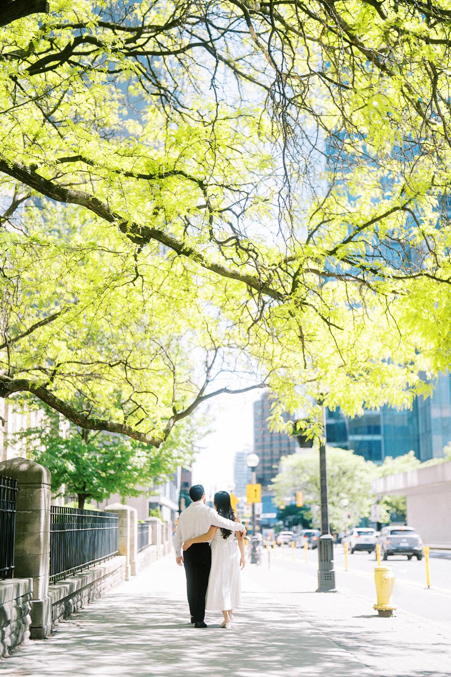 Cassie and Gabriel walk together on a bright city sidewalk in downtown Toronto, with sunlight filtering through green tree branches overhead.