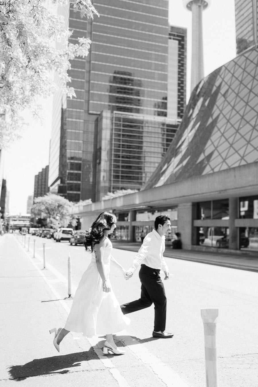 Cassie and Gabriel holding hands as they cross a downtown Toronto street, with city buildings and the CN Tower visible behind them.