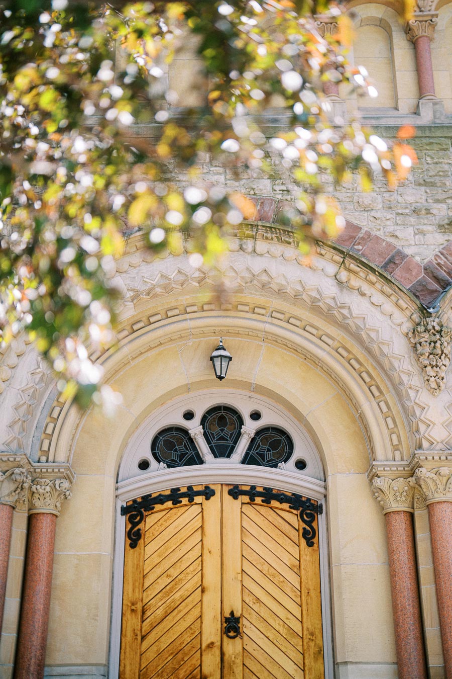 Ornate wooden double doors with ironwork and arched stone entryway of a historic church, partially framed by sunlit tree branches in Toronto.