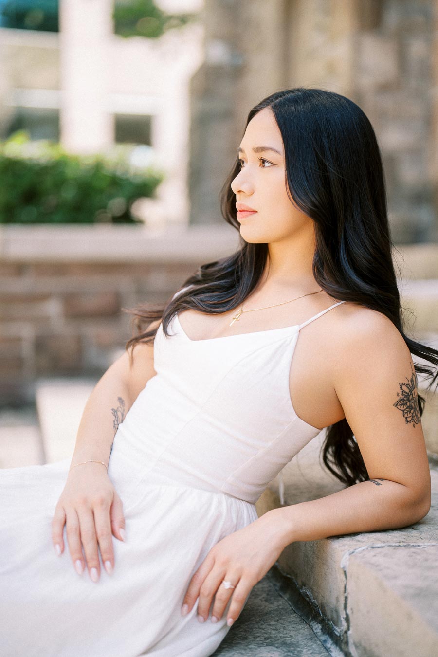 Cassie in a white dress sitting on stone steps, gazing thoughtfully to the side during her engagement session in downtown Toronto.