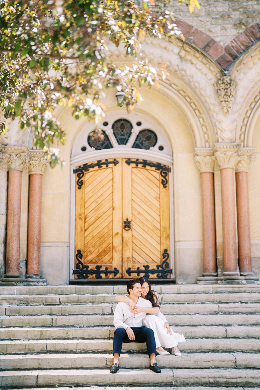 Cassie and Gabriel sit together on stone steps, embracing in front of ornate wooden church doors in downtown Toronto.