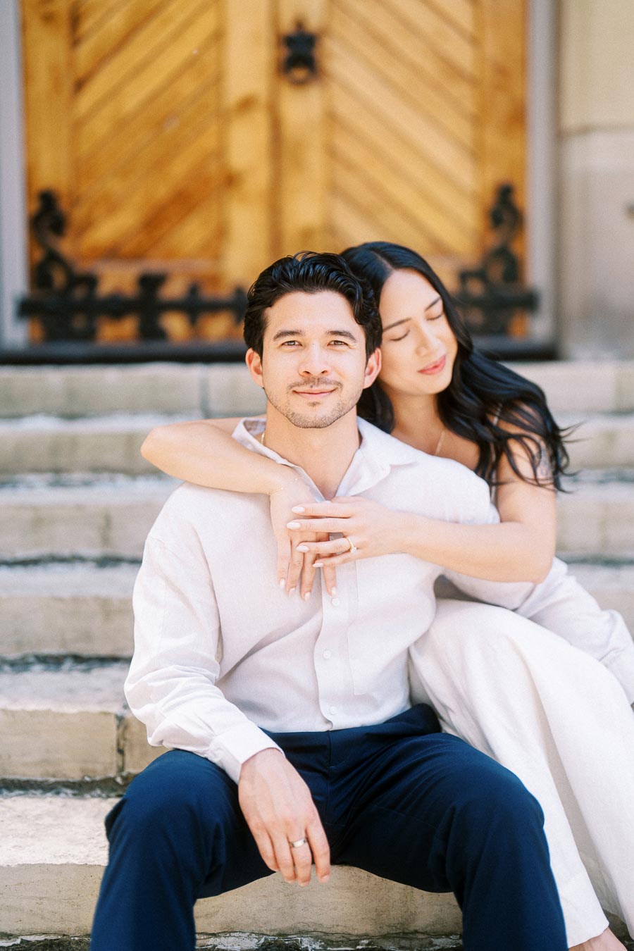 Cassie and Gabriel sit closely together on stone steps in downtown Toronto, with Cassie embracing Gabriel and wooden doors behind them.