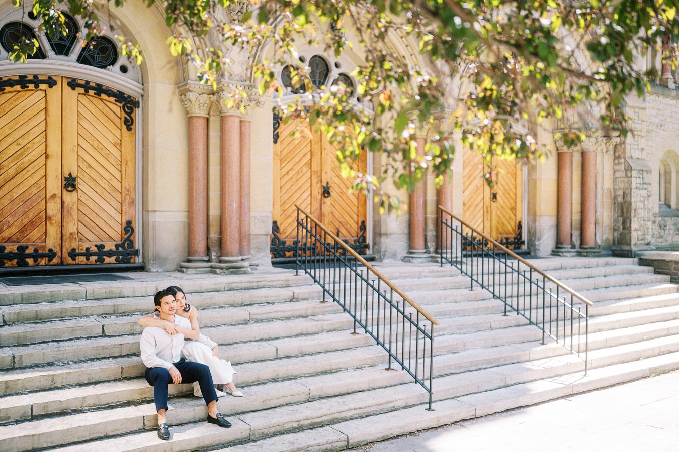 Cassie and Gabriel sit on wide stone steps in front of a historic church with large wooden doors in downtown Toronto.
