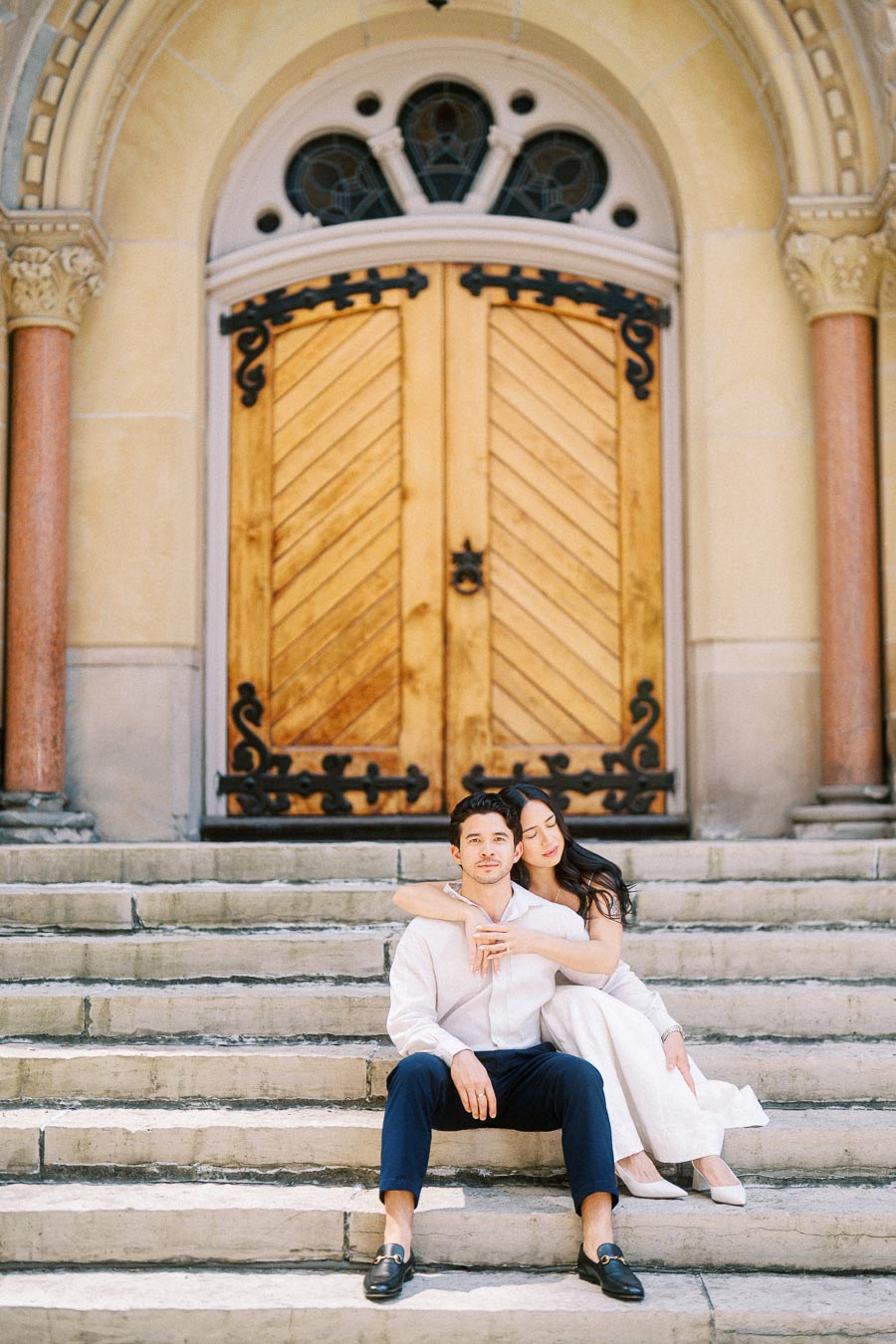 Cassie and Gabriel seated on stone steps in front of large wooden doors during their engagement photoshoot in downtown Toronto.