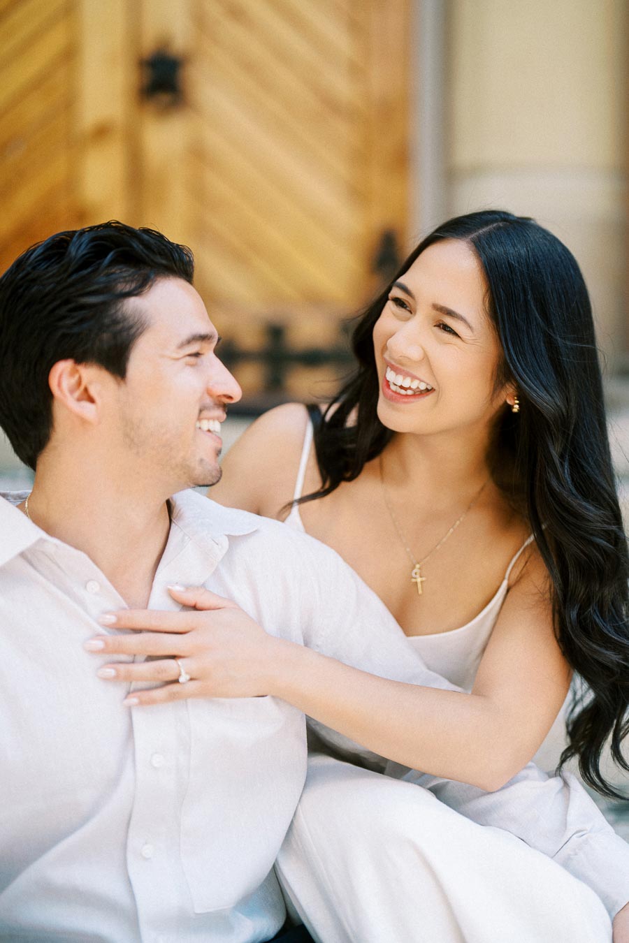 Cassie and Gabriel smiling and laughing together during their engagement photoshoot, seated in front of large wooden doors in downtown Toronto.