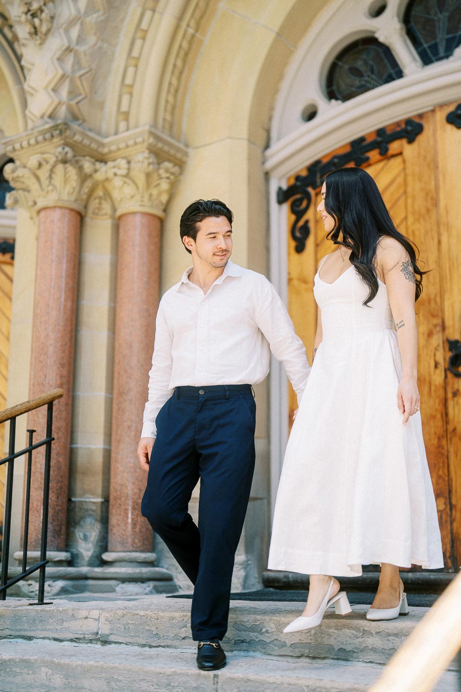 Cassie and Gabriel holding hands and walking down the steps of a historic church in downtown Toronto during their engagement session.