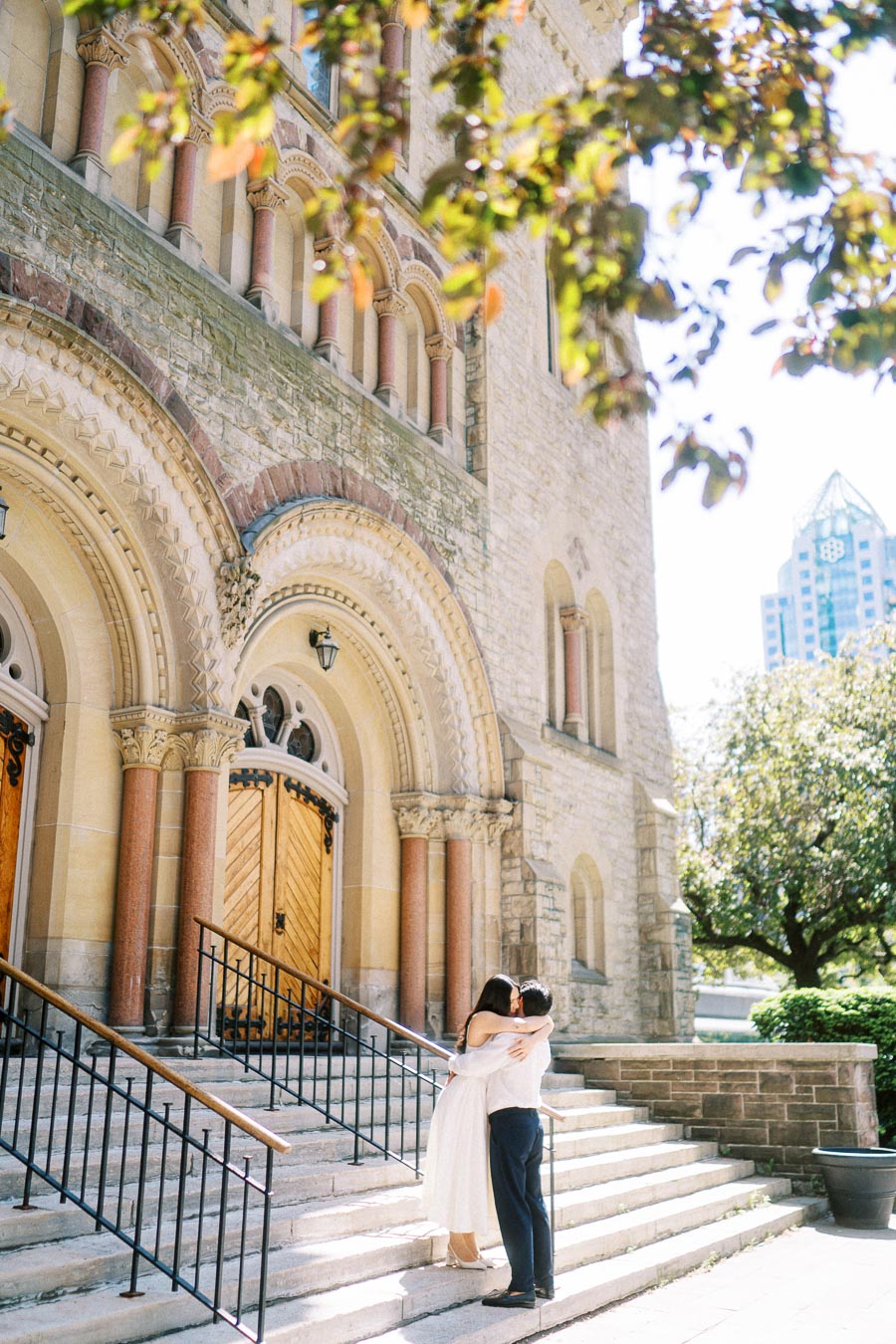 Cassie and Gabriel embracing on the steps of a historic stone church in downtown Toronto, surrounded by leafy trees and city buildings.