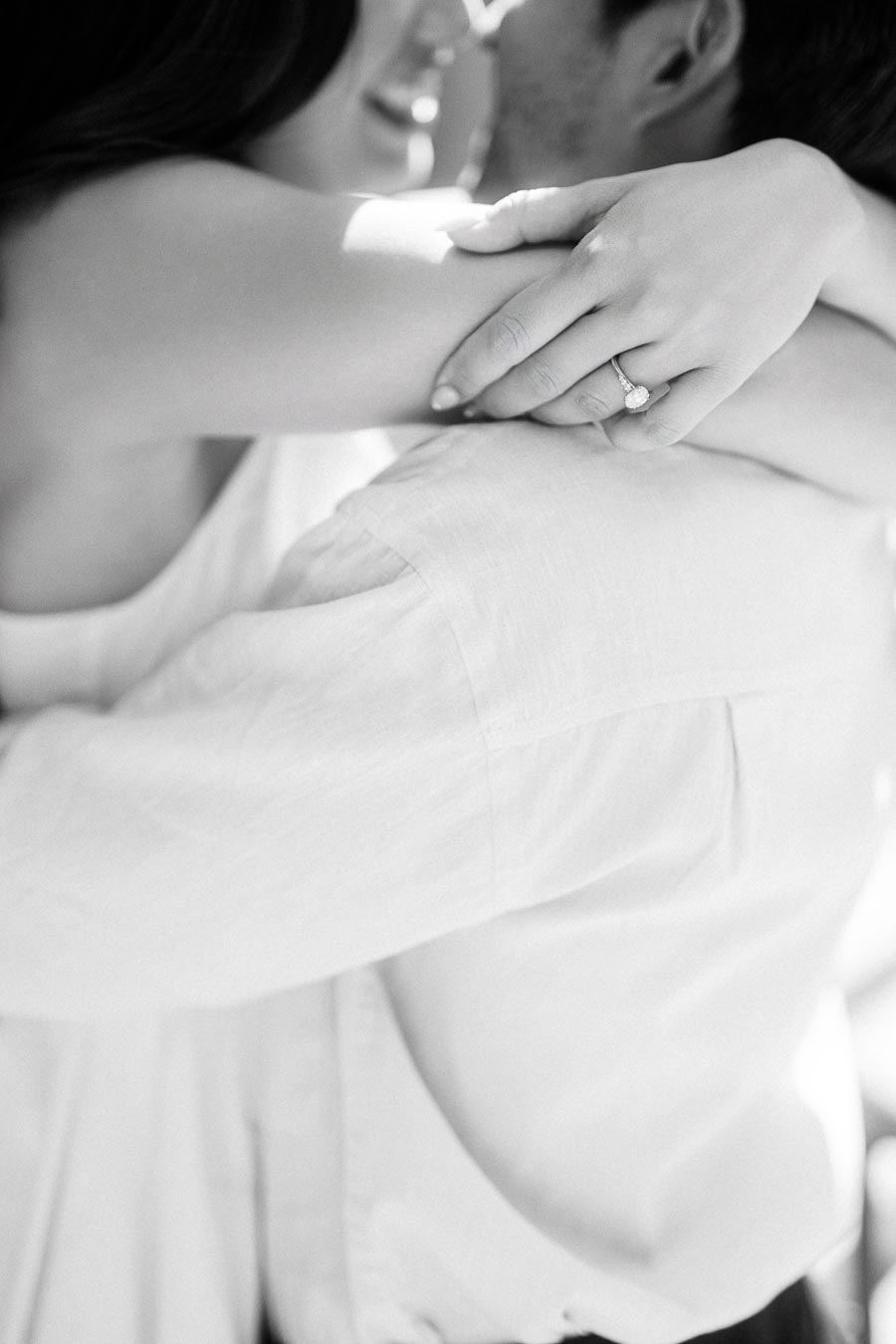 Cassie and Gabriel embrace closely, her hand with an engagement ring resting on his shoulder, both smiling in a black and white photo.