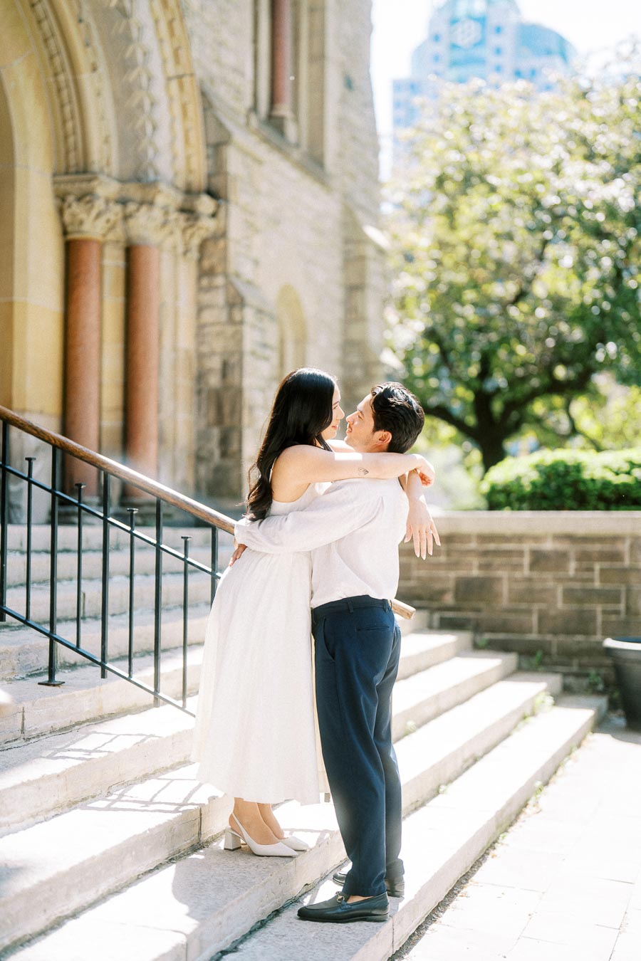 Cassie and Gabriel embracing on the stone steps outside a historic church in downtown Toronto, surrounded by sunlight and greenery.