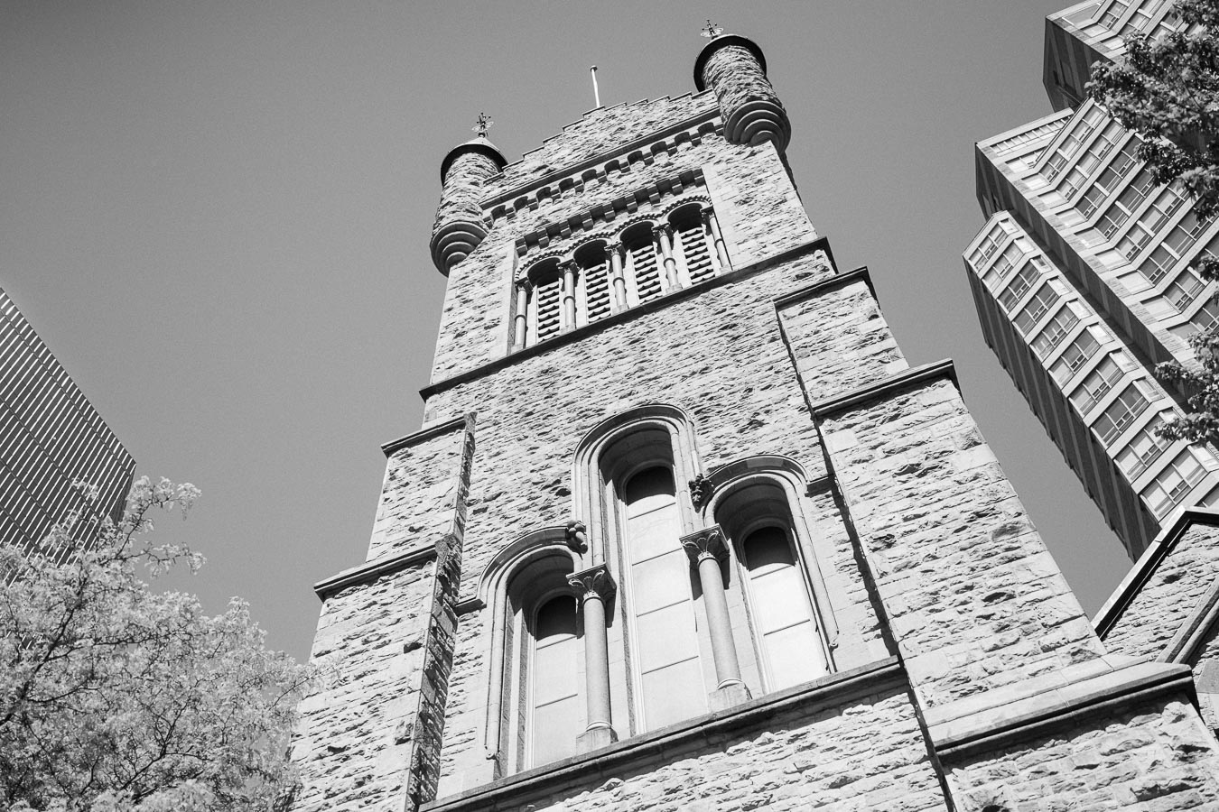 Black and white photo of a historic stone church tower with arched windows, framed by modern office buildings in downtown Toronto.