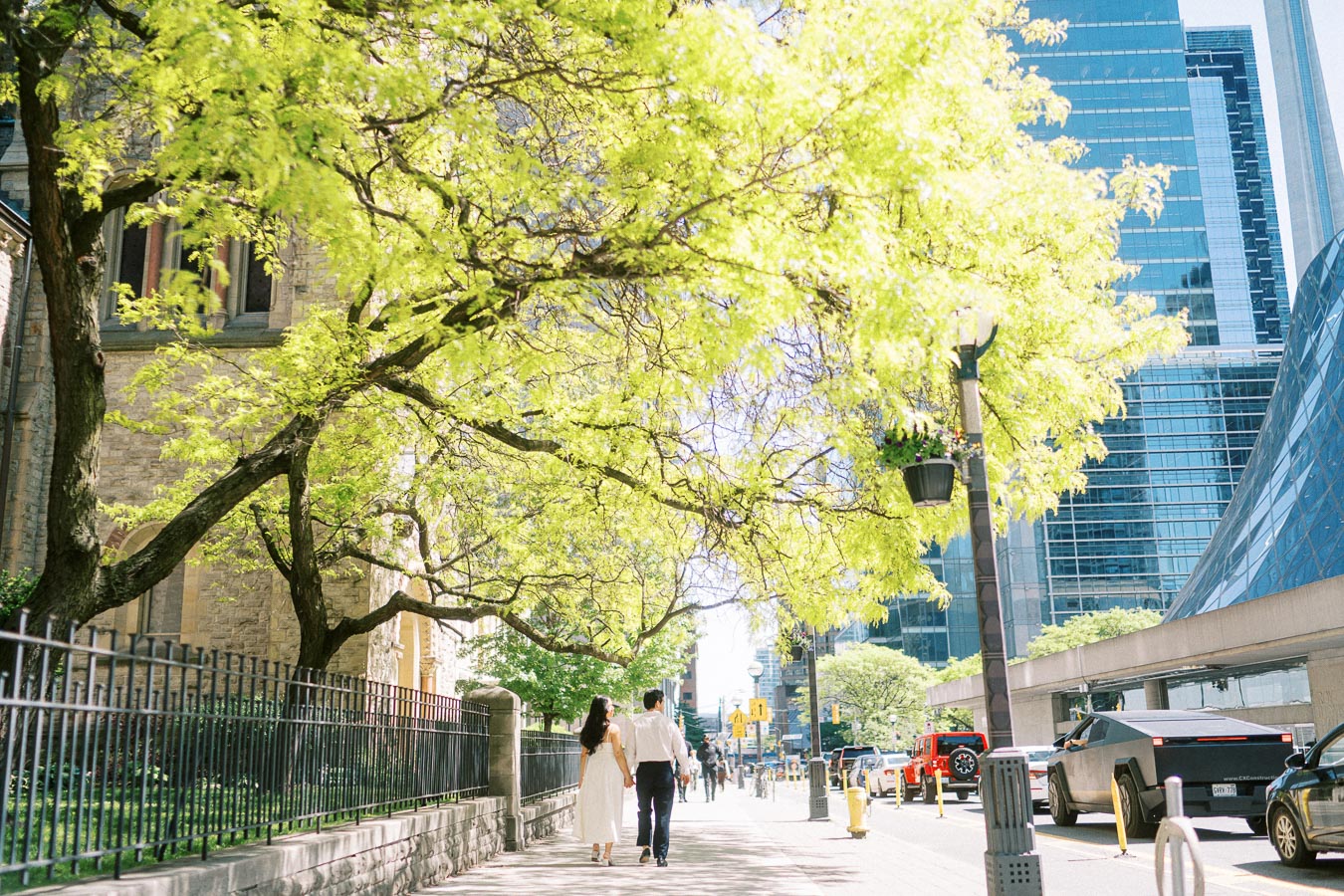 Cassie and Gabriel walk together on a downtown Toronto sidewalk, shaded by leafy trees with urban buildings and cars in the background.