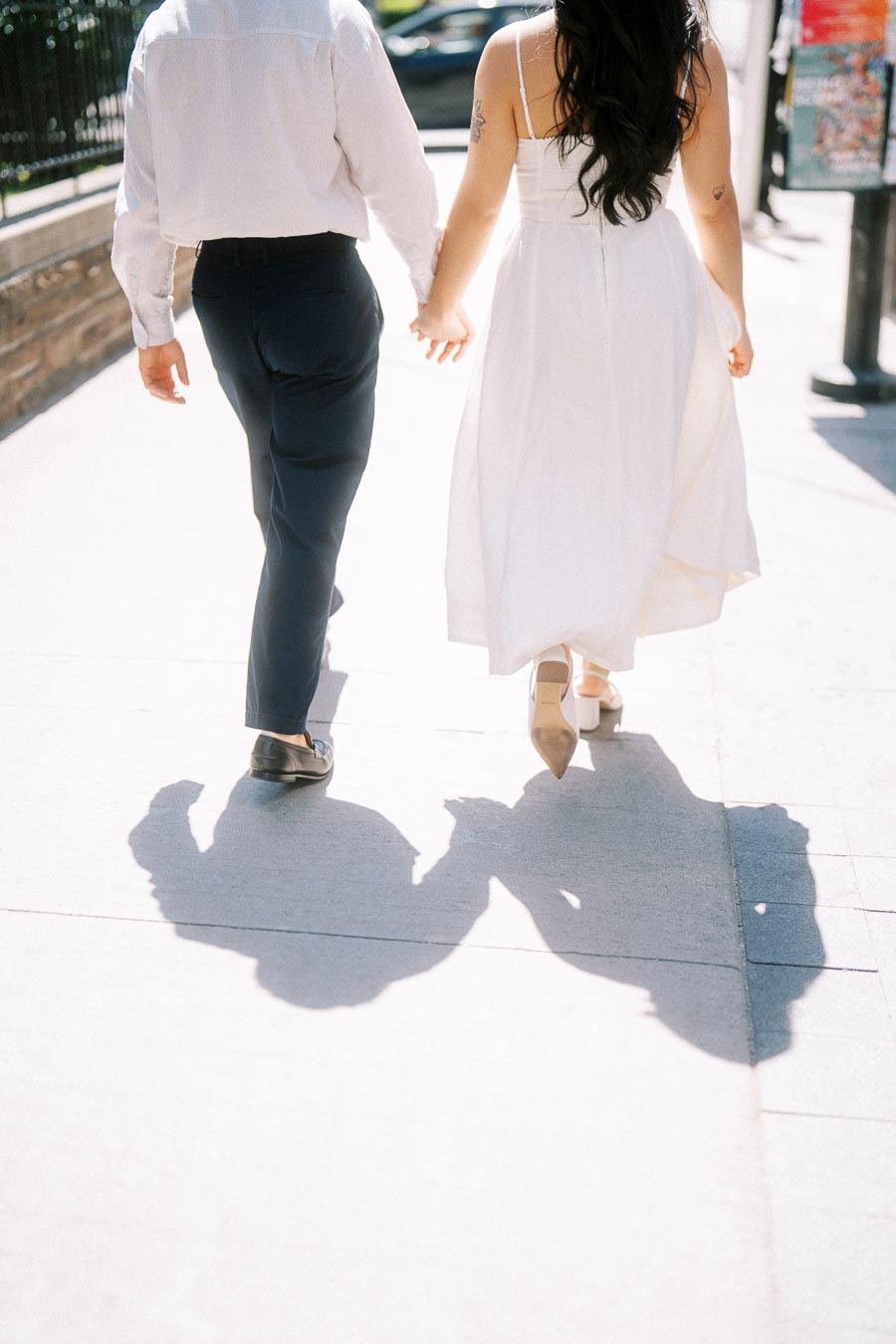 Cassie and Gabriel holding hands while walking on a bright sidewalk in downtown Toronto, casting long shadows on the pavement.