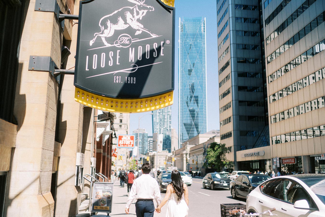 Cassie and Gabriel holding hands as they walk down a busy downtown Toronto street, with tall buildings and The Loose Moose sign visible.