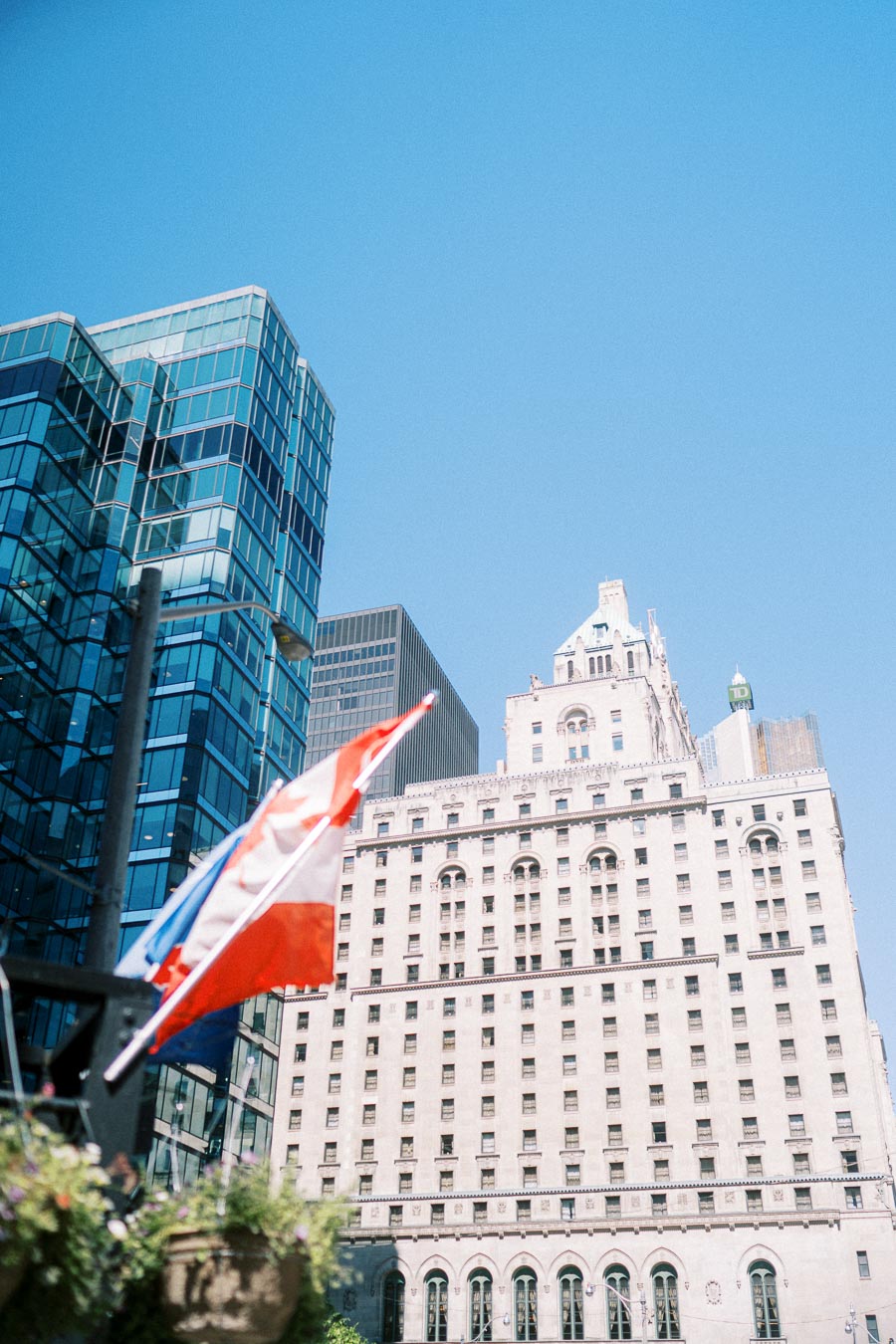Canadian flag and city flags waving in front of a historic stone building and modern glass tower in downtown Toronto on a clear day.
