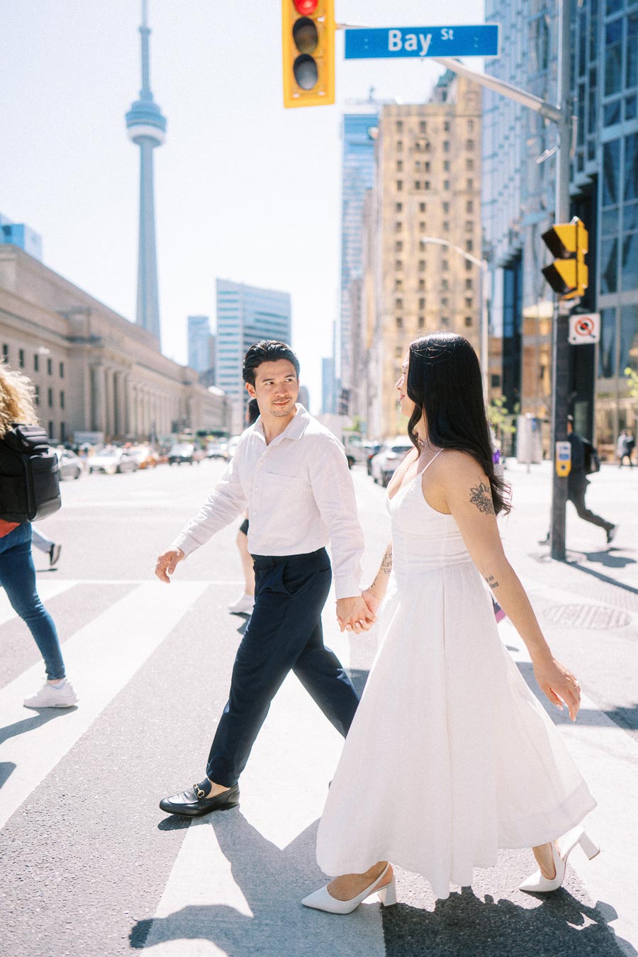 Cassie and Gabriel holding hands while crossing Bay Street in downtown Toronto, with the CN Tower visible in the background.
