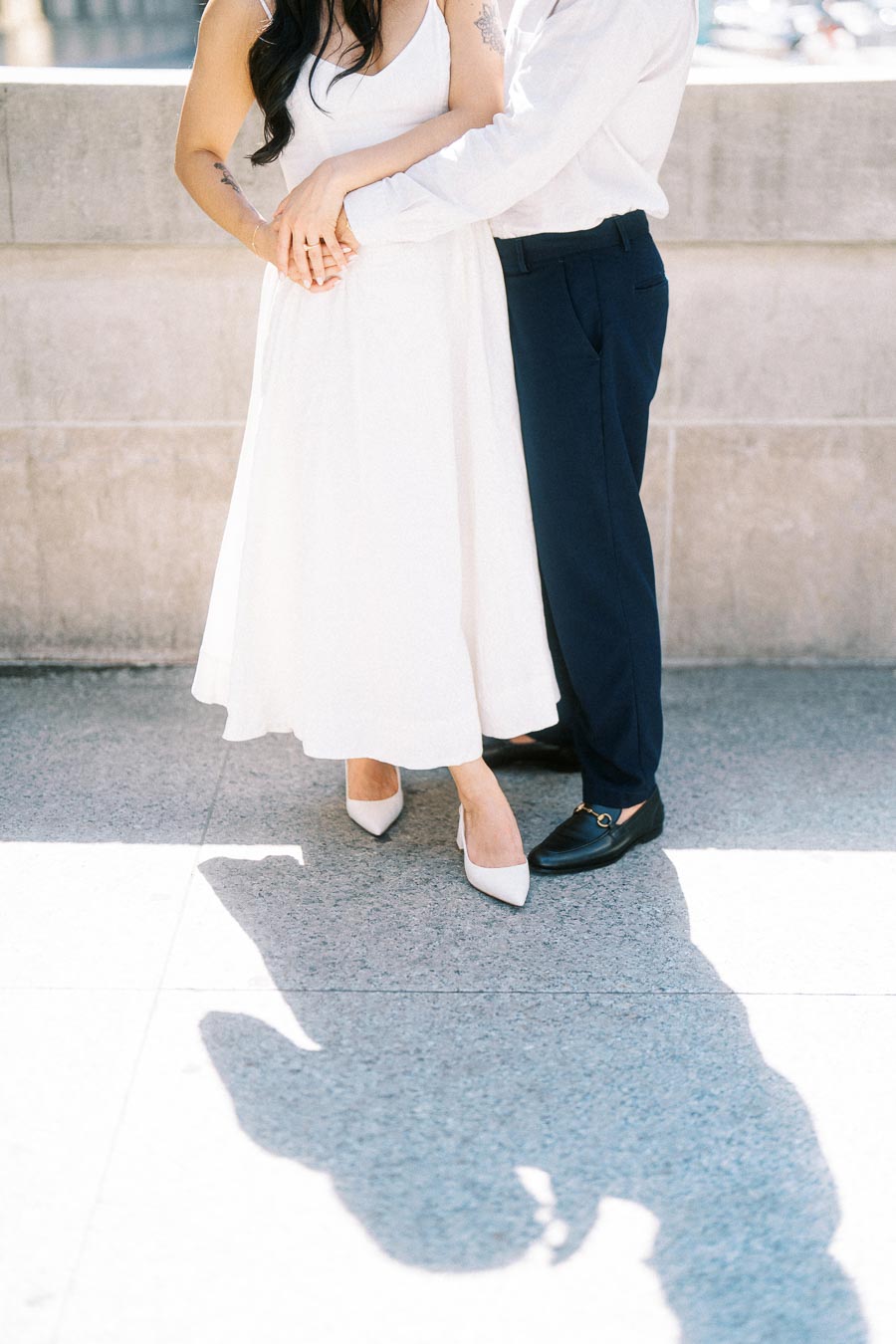 Cassie and Gabriel holding hands, standing together on a sunlit stone walkway in downtown Toronto, only their torsos and legs visible.