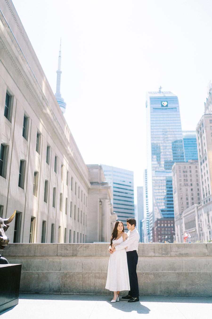 Cassie and Gabriel stand together in an embrace on a downtown Toronto street, with the CN Tower and city skyscrapers visible behind them.