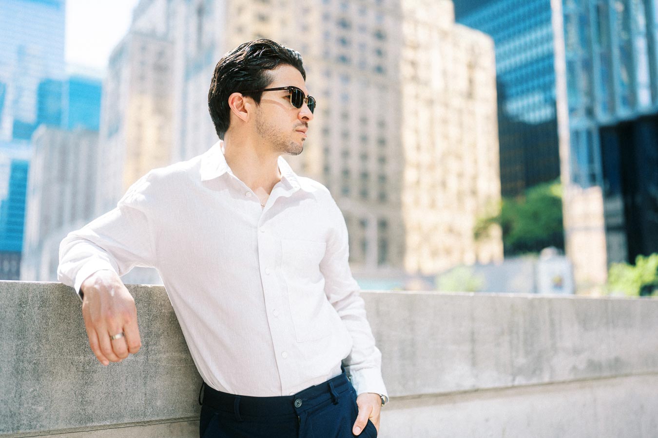 Man wearing a white shirt and sunglasses leaning on a concrete railing with tall office buildings in the background in downtown Toronto.