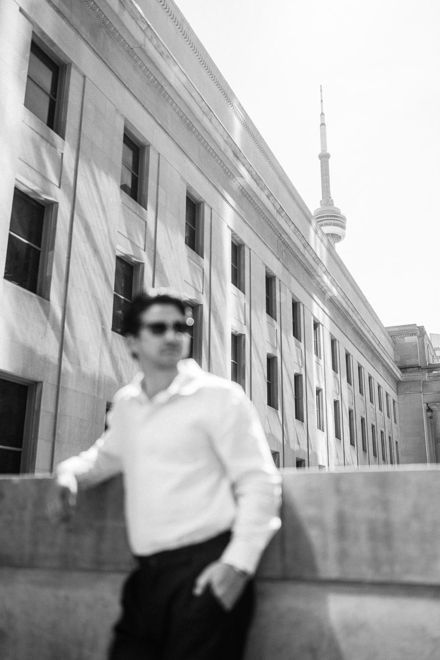 A man in a white shirt stands in front of a stone building in downtown Toronto, with the CN Tower visible in the distance.