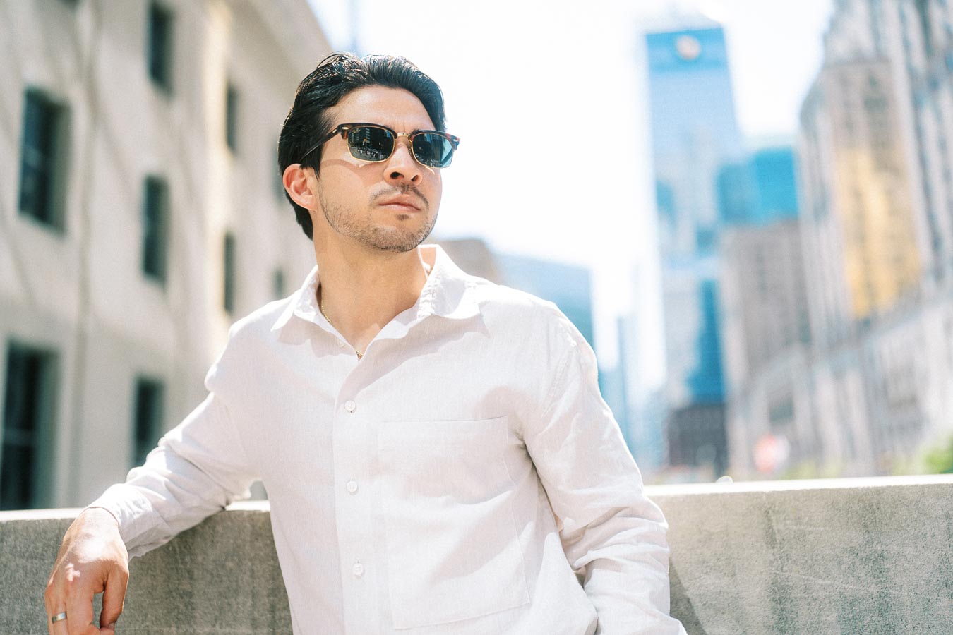 A man wearing sunglasses and a white shirt leans against a stone railing with downtown Toronto's cityscape in the background.