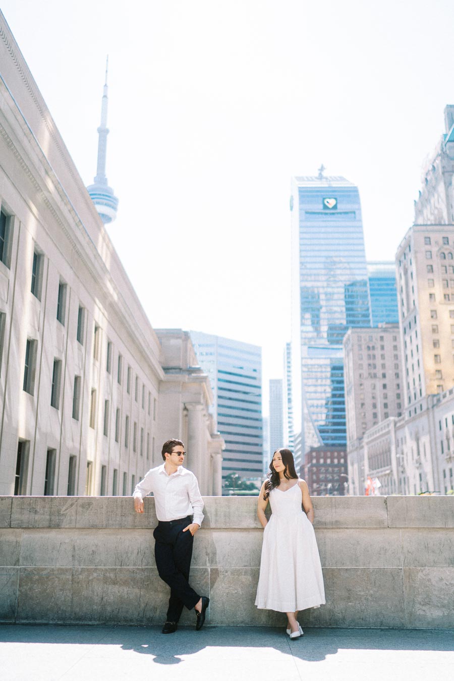 Cassie and Gabriel stand together on a stone terrace in downtown Toronto, with the CN Tower and city buildings visible in the background.