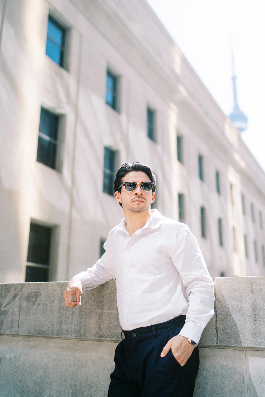 A man in a white shirt and sunglasses leans on a stone ledge in downtown Toronto, with the CN Tower and city buildings in the background.