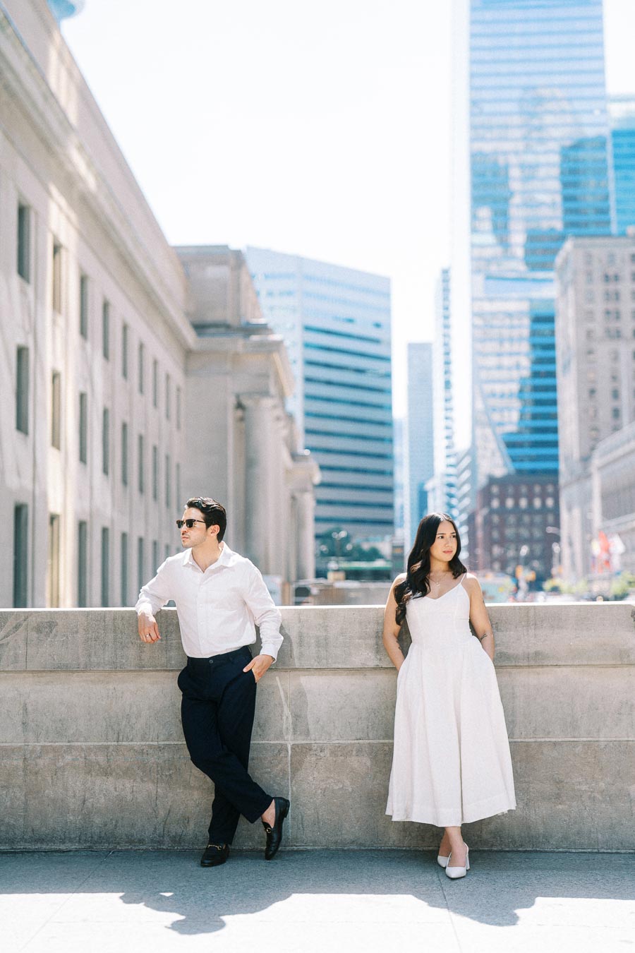 Cassie and Gabriel stand side by side, leaning against a stone wall in downtown Toronto with modern skyscrapers and historic buildings behind them.