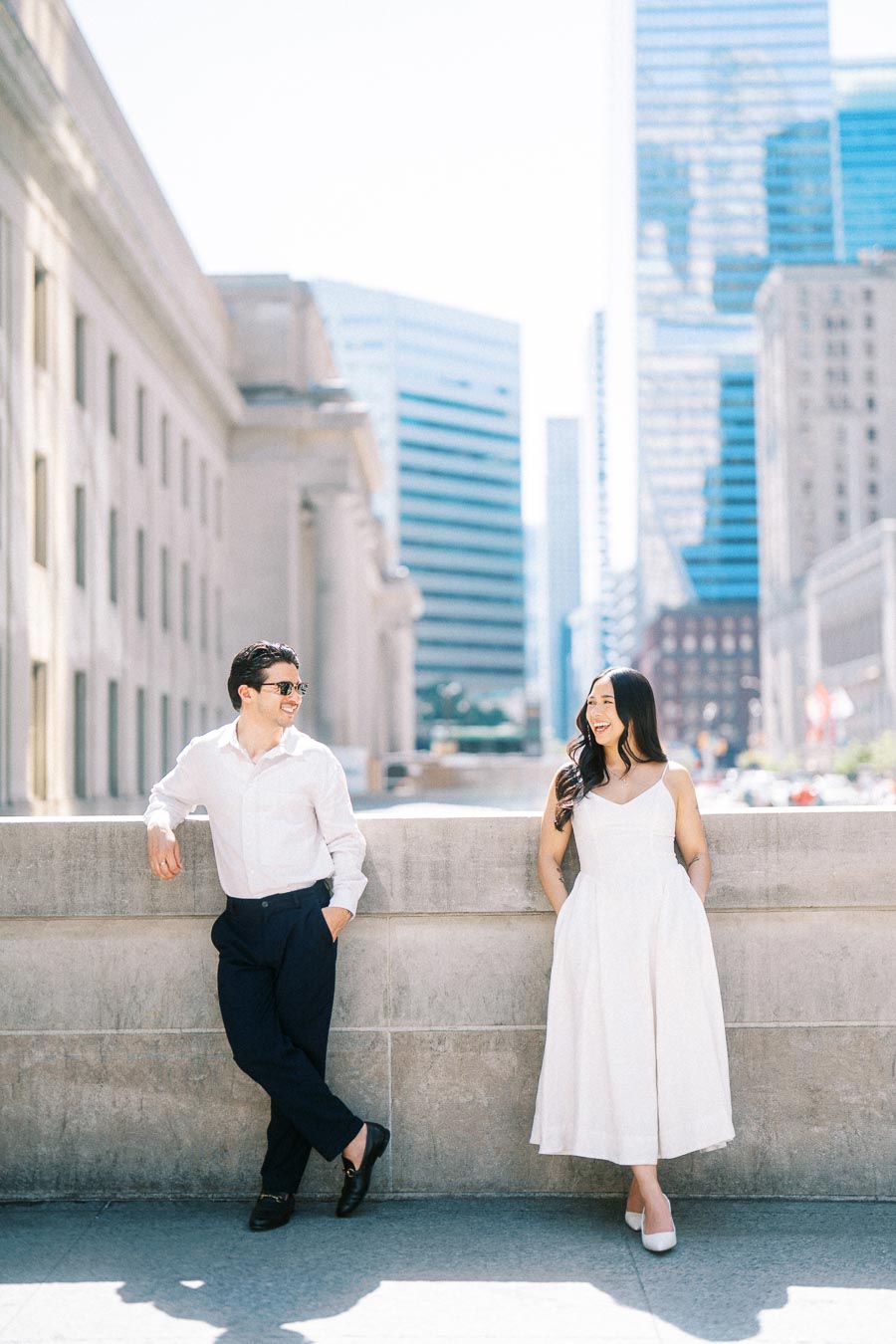 Cassie and Gabriel stand and smile at each other while leaning against a stone wall in downtown Toronto with city buildings in the background.