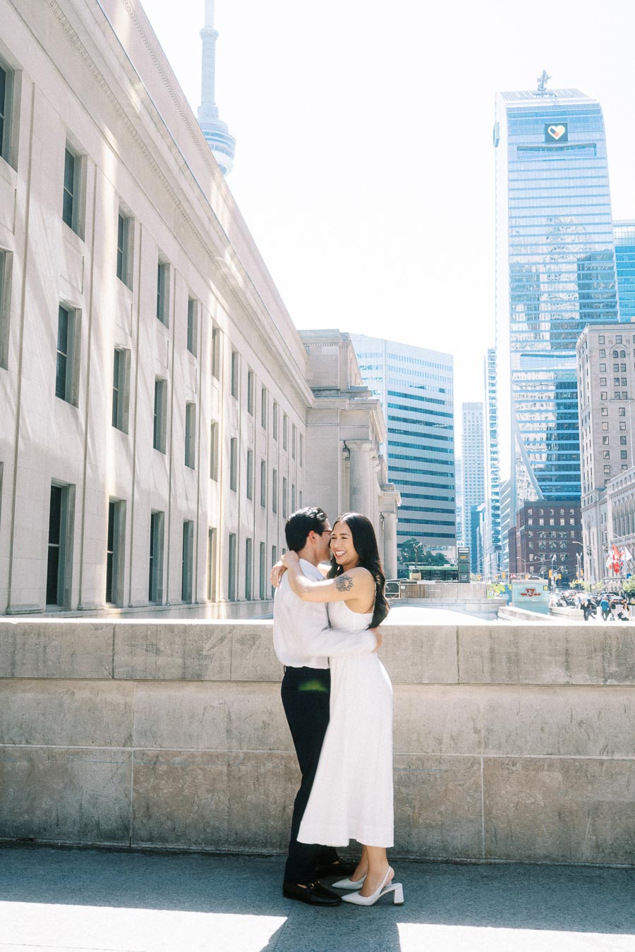 Cassie and Gabriel hugging and smiling outside Union Station in downtown Toronto, with the CN Tower and modern office buildings visible.