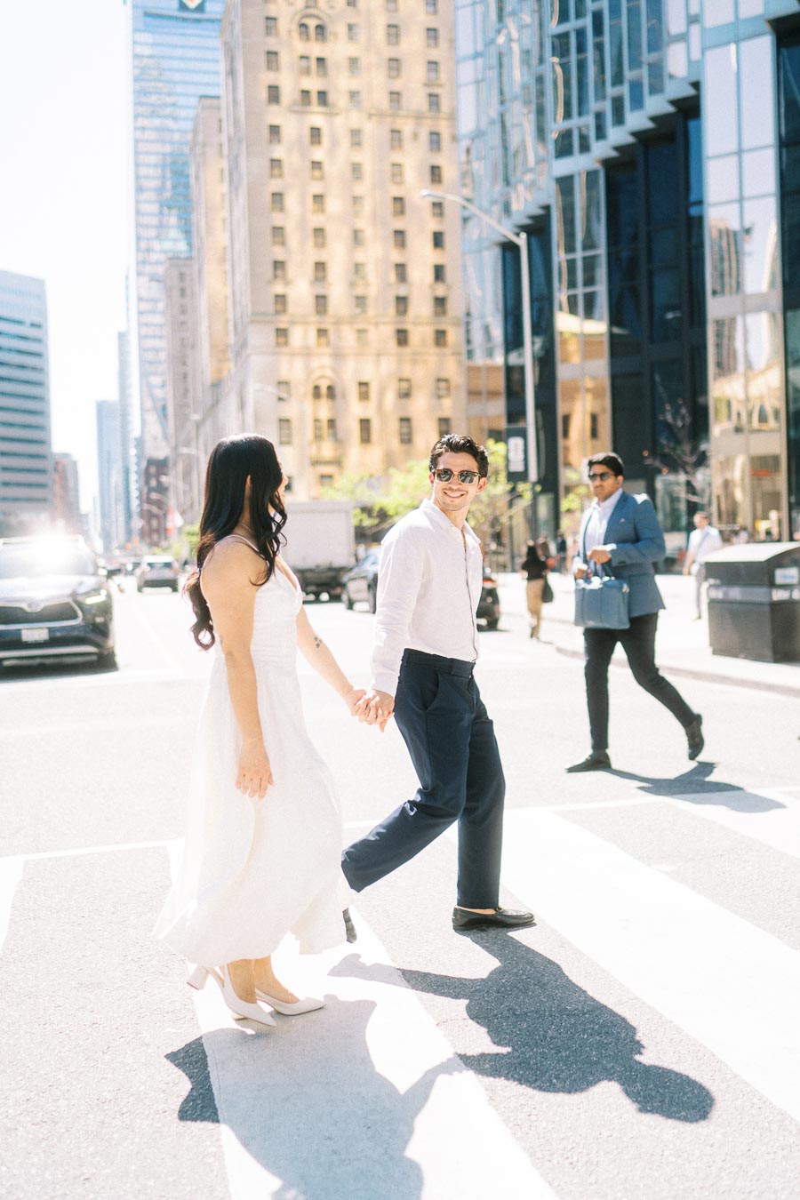 Cassie and Gabriel holding hands and walking across a crosswalk in downtown Toronto with tall buildings in the background.