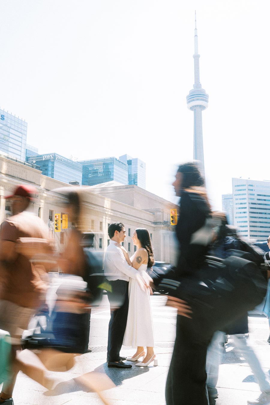 Cassie and Gabriel stand close together on a bustling Toronto street, surrounded by blurred pedestrians, with the CN Tower visible behind them.