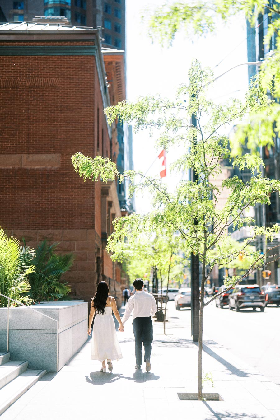 Cassie and Gabriel holding hands while walking away on a bright downtown Toronto sidewalk lined with young trees and urban buildings.