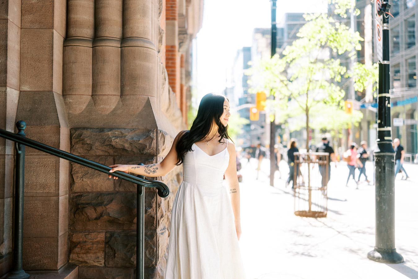 A woman in a white dress stands by stone steps with sunlight streaming onto a busy downtown Toronto street.