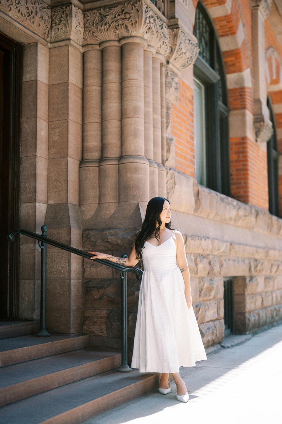 A woman in a white dress stands on stone steps next to an ornate historic building in downtown Toronto, holding a black railing.