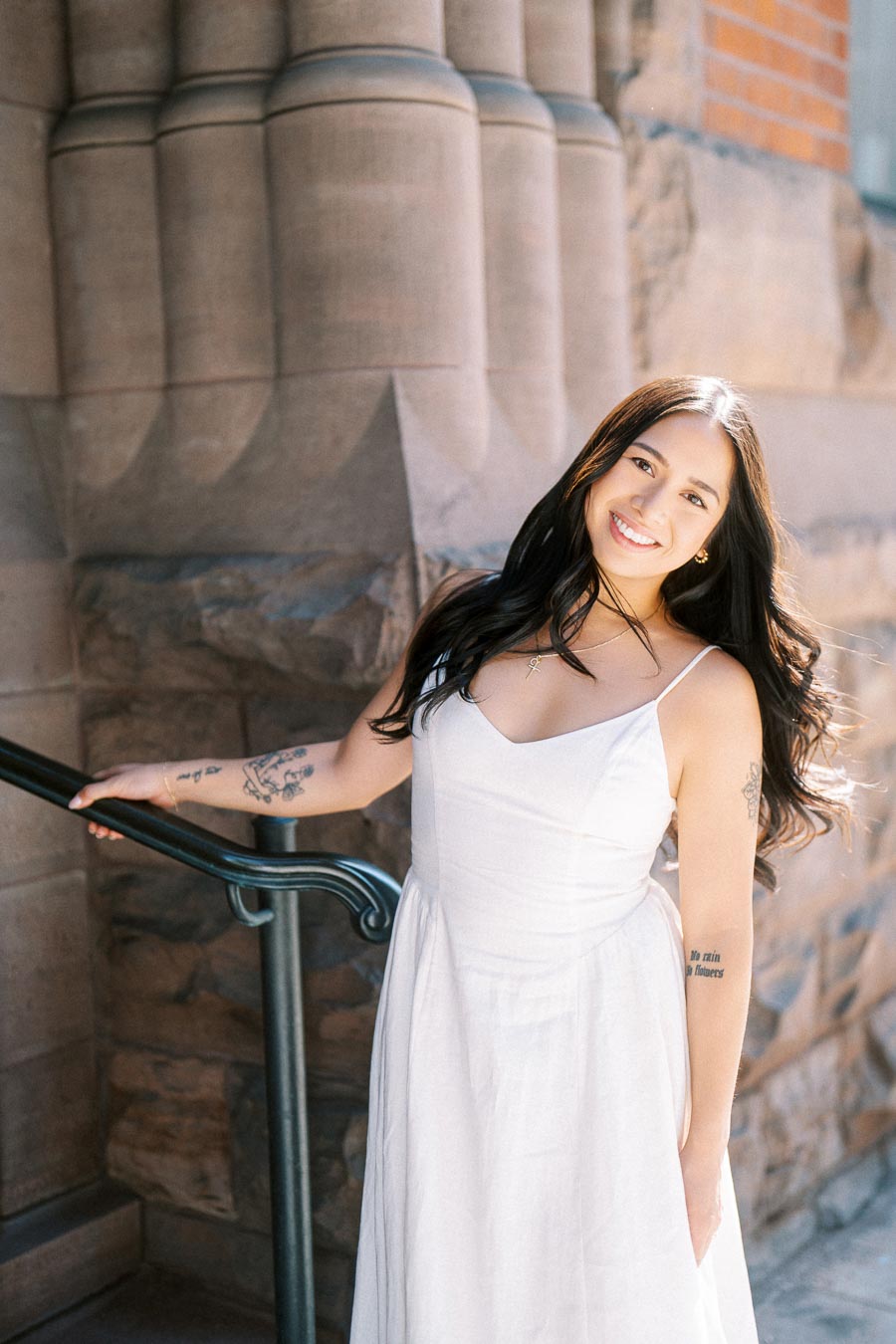 A woman in a white dress stands on stone steps next to a stone wall, smiling at the camera in downtown Toronto.