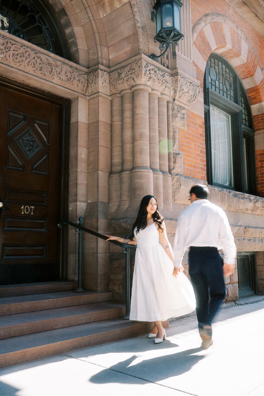 Cassie and Gabriel holding hands on the stone steps of a historic building in downtown Toronto during their engagement session.