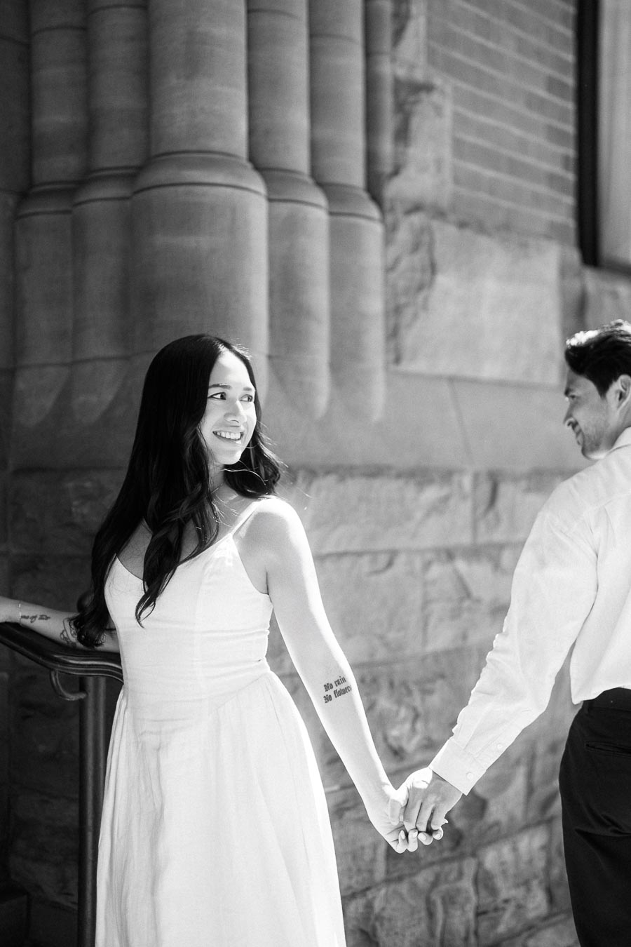 Cassie and Gabriel hold hands while walking near historic stone columns in downtown Toronto during their engagement session.